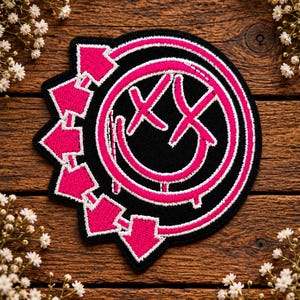 May include: A circular embroidered patch featuring a hot pink smiley face with X-shaped eyes on a black background. The design includes a surrounding ring with five outward-pointing arrows, all outlined in white thread with a distinct stitched border.