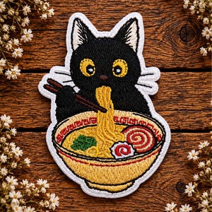 May include: An embroidered patch featuring a black cat with large yellow eyes holding brown chopsticks. The cat is eating yellow noodles from a yellow bowl containing green vegetables and a red and white swirled fish cake, all outlined in white.