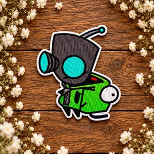 May include: An embroidered patch featuring a stylised character with a dark grey square head, a bright turquoise circular eye, and a single antenna. The body is vibrant green with black accents, a white eye, and a white border outline.
