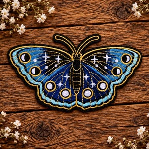May include: An embroidered butterfly patch featuring deep blue and light blue thread with gold outlines. The wings display white star motifs and circular moon phase symbols, including crescents and full circles, set against a dark blue background.
