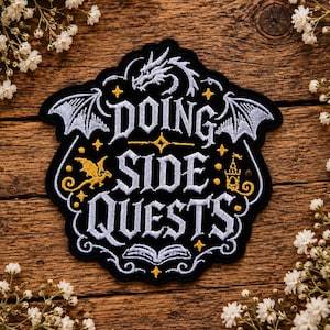 May include: A black embroidered patch featuring the white text DOING SIDE QUESTS. The design includes a white dragon head, wings, an open book, a small gold dragon, a gold castle, and gold stars on a black background with a scalloped border.