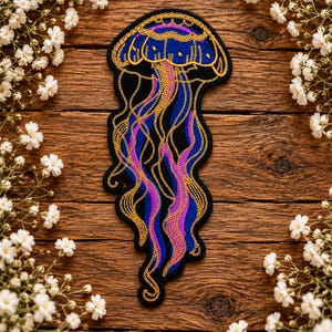 May include: An embroidered patch featuring a jellyfish design with a black background. The jellyfish has a rounded bell outlined in gold thread, with flowing tentacles in shades of deep blue, vibrant purple, and intricate gold metallic stitching.