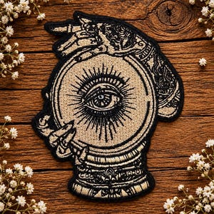 May include: An embroidered patch featuring a black and beige design of a hand resting on a crystal ball. The crystal ball contains a central eye with radiating lines. The hand is adorned with intricate patterns and jewellery, all outlined in black thread.