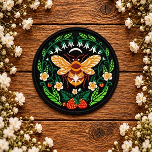 May include: A circular embroidered patch featuring a central bee with yellow and orange wings, surrounded by a wreath of green leaves, white bell-shaped flowers, small white blossoms, and red strawberries on a solid black background.