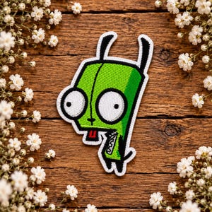 May include: An embroidered patch featuring the character Gir from Invader Zim, depicted in bright green with large white eyes, black ears, and a small red tongue. The patch has a white border and is shaped like the character's head and torso.
