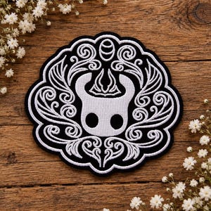 May include: A circular embroidered patch featuring a white stylised character mask with two black circular eyes, centred within an intricate white filigree pattern on a black background, with a scalloped edge border surrounding the entire design.
