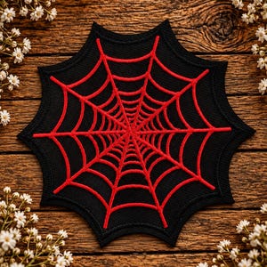 May include: A black fabric patch featuring a vibrant red embroidered spider web design. The patch has a distinct octagonal shape with pointed edges, showcasing detailed stitching that radiates from the centre to the outer perimeter.