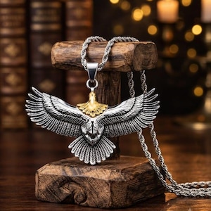 May include: A silver-toned metal pendant shaped like an eagle with wings spread wide, featuring a gold-toned head and neck. The pendant hangs from a silver-toned twisted rope chain, showcasing detailed feather textures across the wings and tail.
