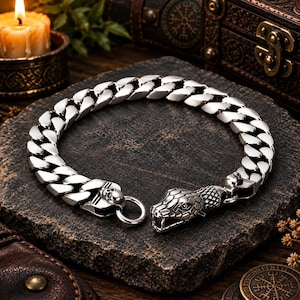 May include: A silver-toned curb chain bracelet featuring a detailed snake head clasp at one end and a small skull accent near the circular ring closure. The metal links are polished and reflective, creating a bold, heavy-duty appearance.