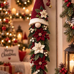 May include: A crochet gnome wall hanging featuring a tall dark red hat with a gold bell, a cream-colored brim, and a round nose. The long beard is composed of dark green crocheted foliage, red and white poinsettia flowers, and small white berries.