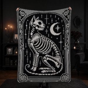 May include: A rectangular black and white woven blanket featuring a detailed pixelated graphic of a cat skeleton in profile. The design includes a crescent moon and star motifs surrounded by an ornate geometric border with spiral corner accents.