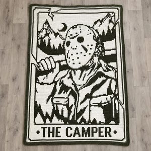 Op de afbeelding: Een rechthoekige gehaakte deken met een zwart-wit grafisch ontwerp van een figuur met een hockeymasker in een bosrijke omgeving. De afbeelding is ingelijst als een tarotkaart met de tekst THE CAMPER in dikke letters onderaan.