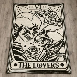 Può includere: Una coperta rettangolare all'uncinetto con un design di carta dei tarocchi in bianco e nero. L'immagine raffigura due teschi rivolti l'uno verso l'altro, circondati da rose. Il bordo superiore mostra il numero romano VI e il basso THE LOVERS.
