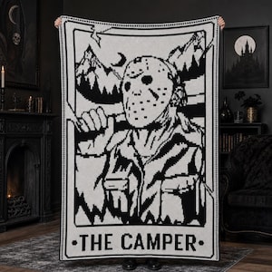 Op de afbeelding: Een rechthoekige gehaakte deken met een zwart-wit grafisch ontwerp van een figuur met een hockeymasker in een bosrijke omgeving. Het ontwerp is ingelijst als een tarotkaart met de tekst THE CAMPER in vette hoofdletters aan de onderkant.