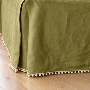 May include: An olive green bed skirt featuring a pleated design and a decorative trim along the bottom edge. The trim consists of a series of small, round, cream-colored pom-poms attached to a fabric border that lines the base of the bed skirt.