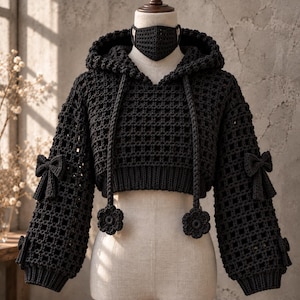 May include: A black crochet hooded sweater featuring a grid pattern, long sleeves with decorative bows, and a matching crochet face mask. The sweater has a ribbed hem and two long drawstrings ending in flower-shaped crochet accents.