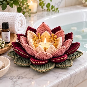 May include: A crocheted lotus flower candle holder featuring layered petals in shades of deep burgundy, soft pink, and cream. The center holds a clear glass votive with a lit candle, surrounded by yellow stamens. The base has sage green leaves.
