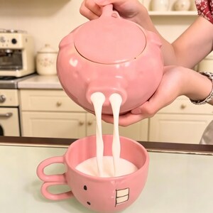 Könnte beinhalten: Eine rosa Keramik-Teekanne in Schweineform, aus der eine weiße Flüssigkeit in eine passende rosa Keramiktasse gegossen wird. Die Tasse zeigt ein Cartoon-Gesicht mit zwei kleinen schwarzen Augen und zwei weißen Zähnen sowie einen Henkel mit zwei Schlaufen.