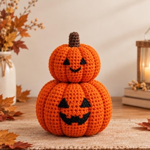 Könnte beinhalten: Eine gehäkelte Kürbisdekoration bestehend aus zwei gestapelten orangefarbenen Kürbissen mit aufgestickten schwarzen Jack-o'-Lantern-Gesichtern. Der obere Kürbis hat einen kleinen braunen Stiel und eine strukturierte Maschenoptik.