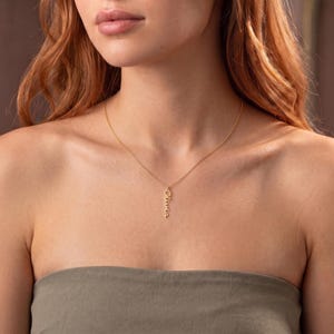 Könnte beinhalten: Eine goldfarbene Halskette mit einer feinen Kette und einem vertikalen Anhänger, auf dem der Name Olivia in Schreibschrift zu lesen ist. Der Anhänger hängt mittig an der Kette, die am Schlüsselbein aufliegt.