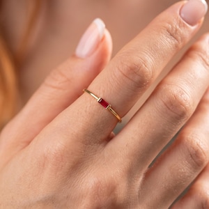 Könnte beinhalten: Ein dünner Goldring mit einem einzelnen, kleinen, rechteckig geschliffenen roten Edelstein, der horizontal in der Mitte gefasst ist. Das Metall hat eine polierte Oberfläche und der Stein wird von zwei kleinen Krappen an den Seiten gehalten.