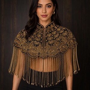 May include: A black sheer capelet featuring intricate gold beadwork in a floral pattern, a high collar, and long gold chain fringe trim that hangs from the scalloped hemline. The garment is open at the front and covers the shoulders and upper chest.