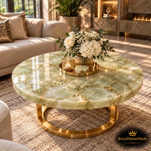 Puede incluir: Una mesa de centro redonda con una superficie de piedra verde claro pulida con vetas naturales y una base circular de metal dorado. Sobre la mesa hay una bandeja dorada con un jarrón de cristal con flores blancas y un pequeño portavelas dorado.
