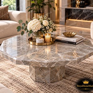 Pode incluir: Uma mesa de centro octogonal feita de mármore castanho polido com veios brancos, com uma base central. Na superfície, encontra-se um tabuleiro circular dourado com um vaso de vidro com flores brancas, um pequeno recipiente e uma vela.