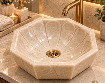 Puede incluir: Un lavabo de mármol beige con un diseño interior facetado en forma de pétalos y un desagüe central de latón. El lavabo está colocado sobre una encimera de mármol a juego bajo un grifo de latón alto y curvado.