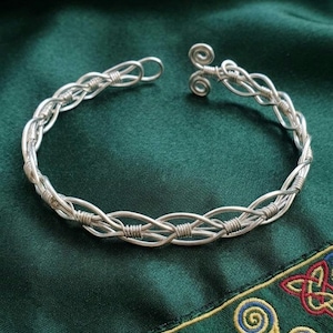 May include: A silver-coloured wire cuff bracelet featuring a braided design with multiple strands intertwined and secured by small wire wraps. The bracelet has an open-ended construction with two small, circular spiral terminals at each end.