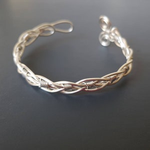 Puede incluir: Un brazalete plateado con un diseño trenzado. El brazalete presenta una serie de hebras entrelazadas, creando una apariencia texturizada. Los extremos del brazalete tienen remolinos decorativos y un cierre de gancho. La joya se muestra sobre un fondo oscuro.