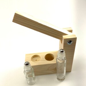 May include: A wooden roller bottle holder with two clear glass roller bottles with silver tops. The holder has a hinged lid that opens to reveal two slots for the bottles.