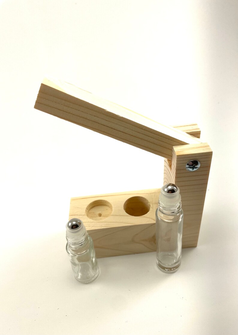 May include: A wooden roller bottle holder with two clear glass roller bottles with silver tops. The holder has a hinged lid that opens to reveal two slots for the bottles.