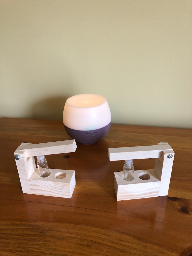 May include: Two wooden holders with small glass bottles inside, next to a white and purple essential oil diffuser on a wooden table.