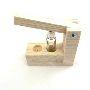 May include: A wooden bottle stopper with a clear glass bottle inside. The stopper is made of light wood and has a simple design. The bottle is held in place by a wooden frame.