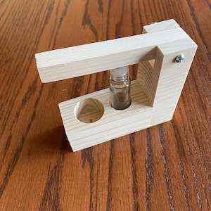 May include: A wooden roller bottle holder with a small glass bottle inside. The holder is made of light wood and has a simple design.