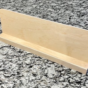 May include: A light-colored wooden shelf with a back panel and a small front lip. The shelf is made of unfinished wood and is designed to hold books or other items. The shelf is approximately 12 inches long.