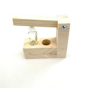 May include: A wooden stand with a small glass bottle in the center. The stand has a hinged top that can be lifted to access the bottle.