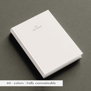 May include: A white hardback planner with the text MY PLANNER printed in black on the front cover and the spine. The planner is positioned at an angle, and a white banner at the bottom displays the text 60+ colours | Fully customisable.