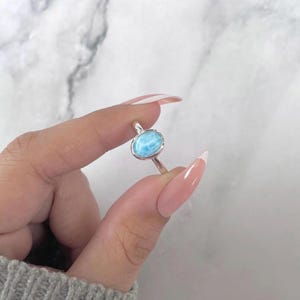 May include: A silver ring featuring a polished, oval-shaped light blue Larimar gemstone set in a smooth metal bezel. The band is thin and metallic, showcasing the natural marbled texture and sky-blue color of the stone in a simple, clean design.
