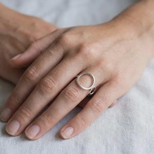 May include: A silver ring featuring a large circular band design with a smooth, matte finish. The ring is constructed from a single continuous piece of metal that forms a prominent open circle, creating a clean and simple aesthetic for the finger.
