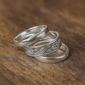 May include: A set of three silver rings stacked together. The top and bottom rings have a smooth, polished finish, while the middle ring features a detailed, embossed floral pattern with leaves and small blossoms encircling the entire band.
