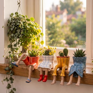 Peut inclure: Quatre cache-pots en céramique en forme de bas du corps assis sur un rebord. Le premier porte un short rouge et des sandales bleues, le deuxième une jupe à carreaux blancs et des bottes rouges, le troisième un pantalon jaune, le quatrième un jean bleu.