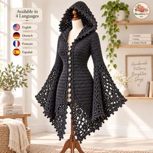 May include: A dark gray crochet hooded cardigan featuring long flared sleeves with intricate lace patterns, a front button closure, and decorative pom-pom trim along the hood and hem edges. The garment is displayed on a mannequin stand.