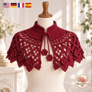 Könnte beinhalten: Ein dunkelrotes Häkel-Capelet mit Stehkragen, Rautenmuster und Blumenmotiven am gewellten Saum. Das Kleidungsstück verfügt über zwei herabhängende Bänder mit kleinen Blumenakzenten an den Enden und wird auf einer Schneiderpuppe präsentiert.