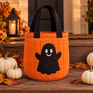 Könnte beinhalten: Eine gesteppte orangefarbene Stofftasche in Eimerform mit einer schwarzen Geisterapplikation, die weiße Augen und einen lächelnden Mund aufweist. Die Tasche hat zwei schwarze Stoffgriffe und vertikale Nähte, die ihr ein gepolstertes Aussehen verleihen.