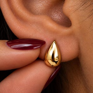 May include: A polished gold stud earring shaped like a smooth, rounded teardrop. The earring features a reflective, high-shine metallic surface with a tapered top and a wider, rounded base, securely positioned against the earlobe.