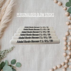 May include: A stack of six off-white cylindrical glow sticks, each printed with the black text Abdul Hanin Hassan 23/05/26. The text PERSONALISED GLOW STICKS is displayed above the sticks, which are arranged in a pyramid formation.