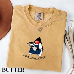 May include: A butter-colored t-shirt with a crew neckline and a Comfort Colors label. The graphic features a stylized character wearing a blue apron, pouring coffee into a cup, with the text JAVA DEVELOPER printed below in black.