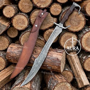 May include: A Damascus steel dagger with a dark wooden handle and a curved metal pommel, accompanied by a brown leather sheath featuring two silver-colored metal rivets and an embossed design. The text AURA CRAFTS WORKS is visible.